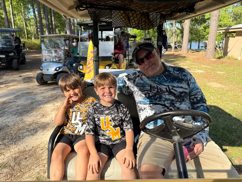 An adult and two smiling children together in a golf cart among the pines